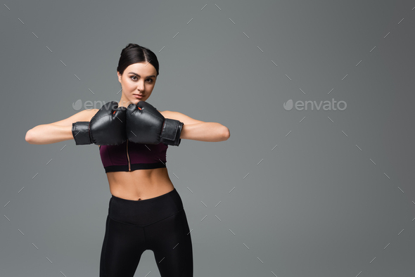 young sportswoman in boxing gloves looking at camera isolated on grey