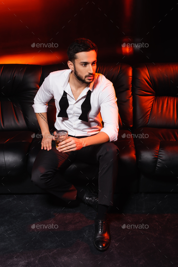 Man in formal wear holding glass of whiskey on couch on black background with red lighting