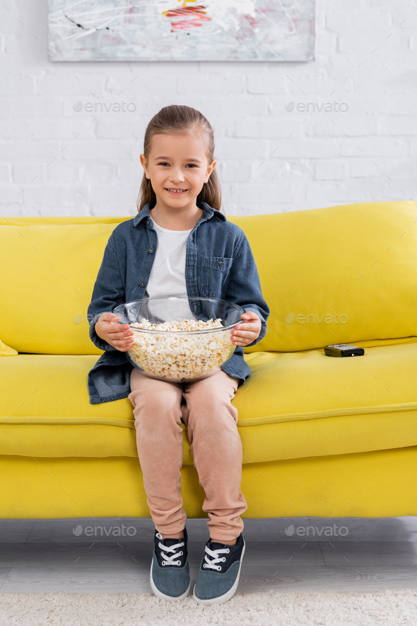 Kid with popcorn smiling at camera near remote controller on couch