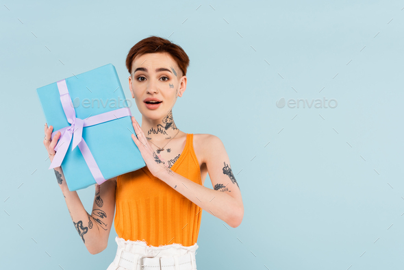 amazed and tattooed woman holding wrapped present isolated on blue