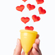 A hand holding a yellow menstrual cup on a white background with pink hearts as menstrual drops. - PhotoDune Item for Sale