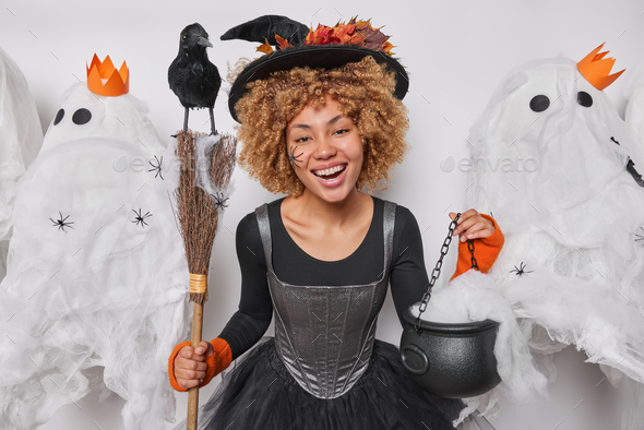 Halloween party celeration. Positive curly haired young woman dressed in black dress and wizard hat
