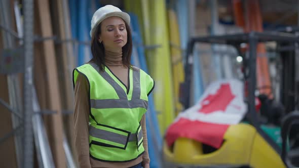 Serious Confident Beautiful Woman in Hard Hat Looking Around at Racks with Goods in Warehouse and alt
