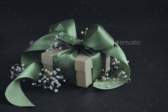 Paper gift box with olive green ribbon tied in a bow, small flowers, black  background. Stock Photo by TaniaJoy