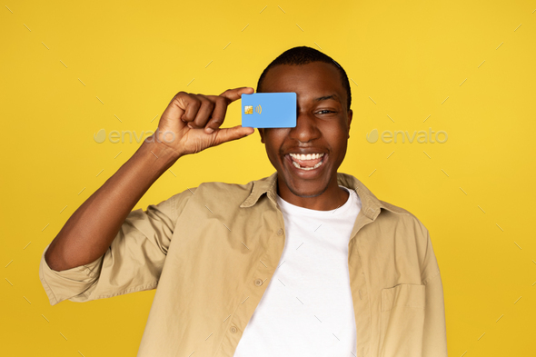 Laughing young african american guy in casual puts credit card to eye, have fun