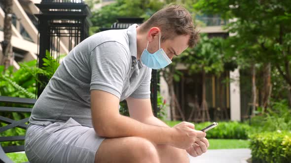 Sad Man Wear Face Mask Sit Hunched Over in Public Park Work with Smartphone alt