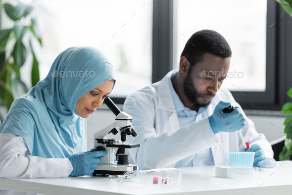 Muslim scientist looking through microscope near blurred african american colleague working in