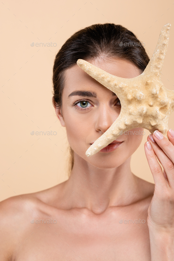 charming woman with naked shoulders obscuring face with starfish isolated on beige, beauty concept