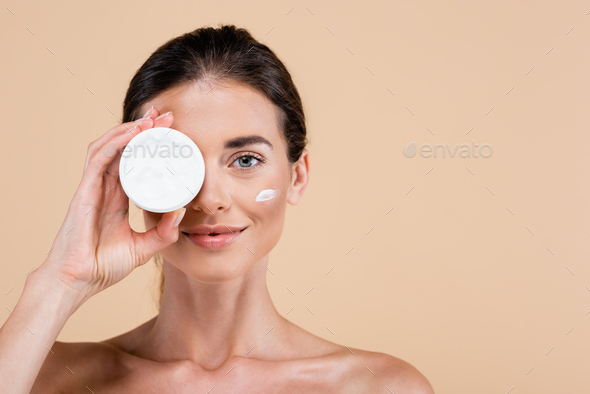 woman with naked shoulders obscuring face with face cream container isolated on beige