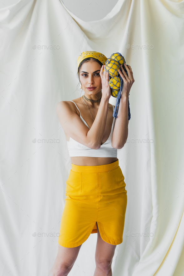 pretty woman in yellow headscarf and crop top holding string bag with lemons on white