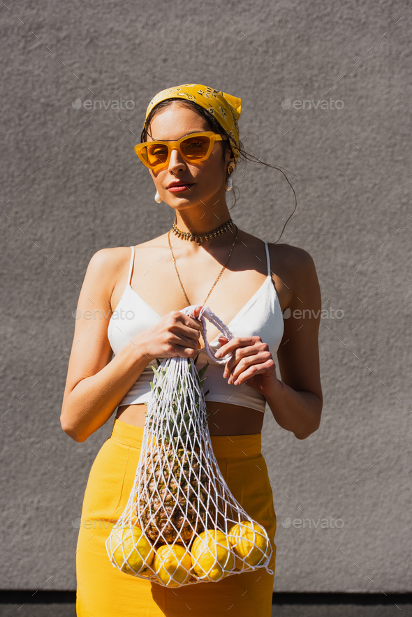 stylish woman in yellow sunglasses and headscarf holding string bag with fresh fruits near concrete