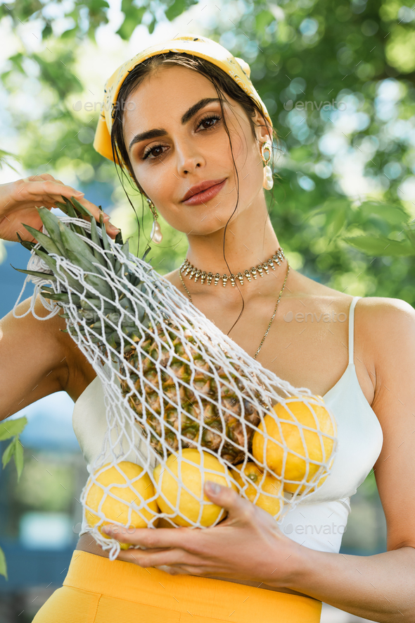 trendy woman in yellow headscarf holding string bag with fruits