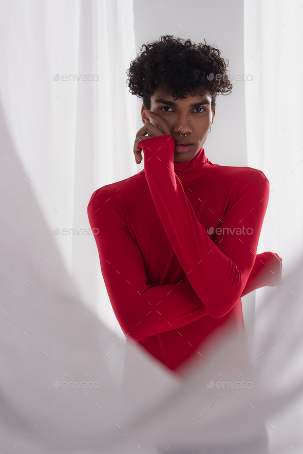 young african american man in red turtleneck posing with hand near face near white tulle cloth
