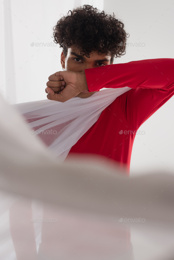 african american man looking at camera while obscuring face with arm near white blurred drapery