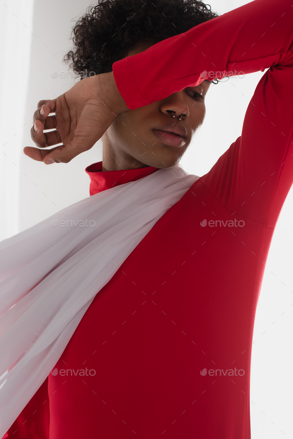 young african american man in red turtleneck covering face with arm while posing near white drapery