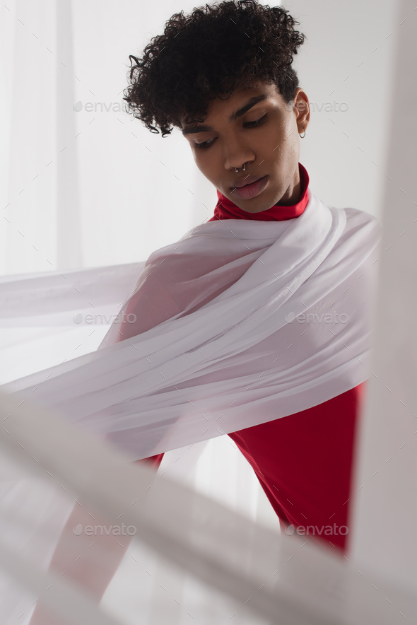 stylish african american man wearing red turtleneck posing in white tulle drapery