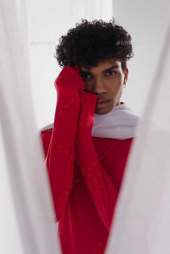 young african american man in red turtleneck posing near white chiffon cloth with hands near face