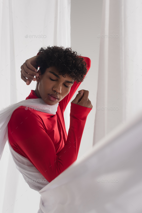 african american man in red turtleneck posing with closed eyes near white tulle drapery