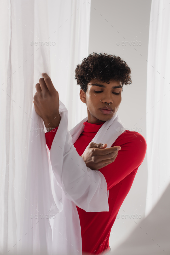 curly african american man in red turtleneck posing near white chiffon cloth