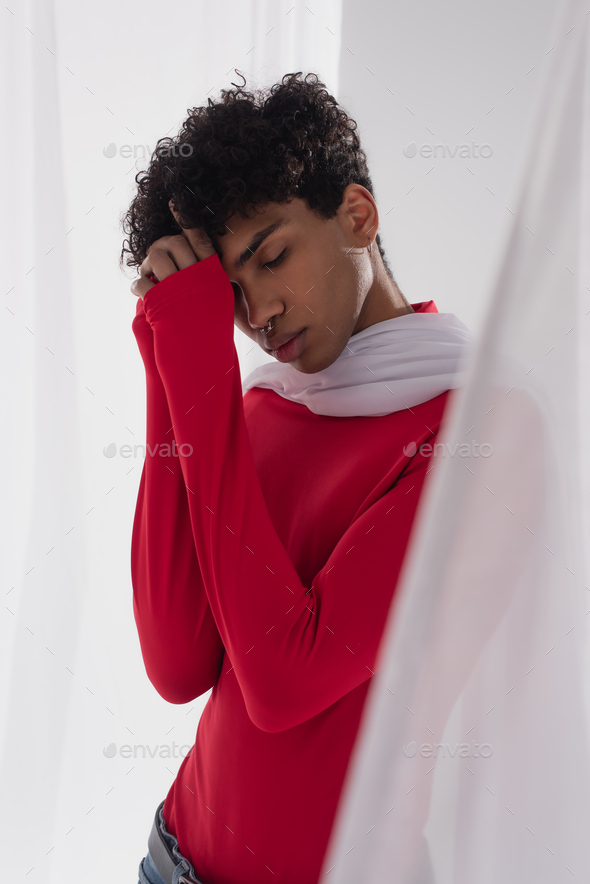 african american man in red turtleneck posing near white tulle cloth with closed eyes and hands near