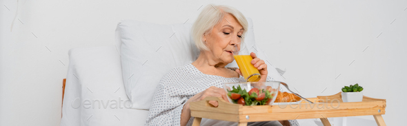 Senior patient drinking orange juice near croissant and salad on tray in hospital, banner
