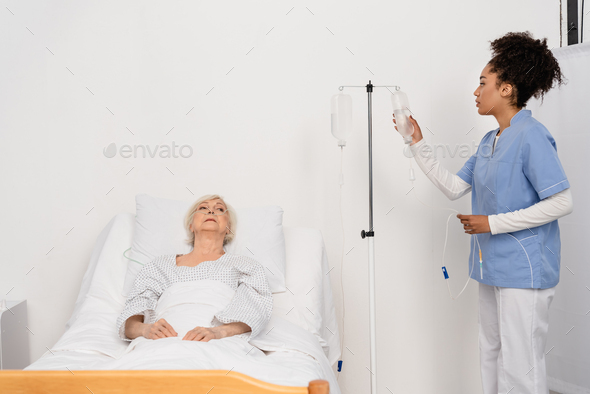 African american nurse holding bottle of intravenous therapy near senior patient in hospital