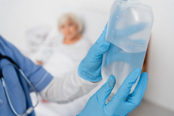 Nurse in latex gloves holding bottle of intravenous therapy near blurred patient