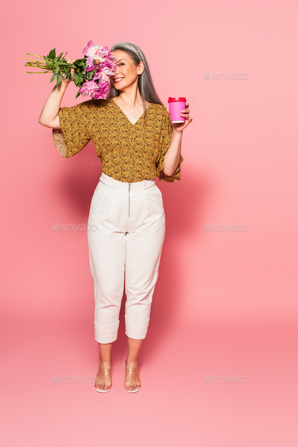 happy asian woman with takeaway drink obscuring face with peonies on pink background