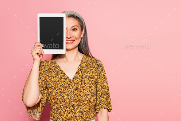 smiling asian woman obscuring face with digital tablet isolated on pink