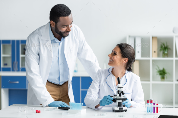 Scientist smiling at african american colleague near microscope and test tubes