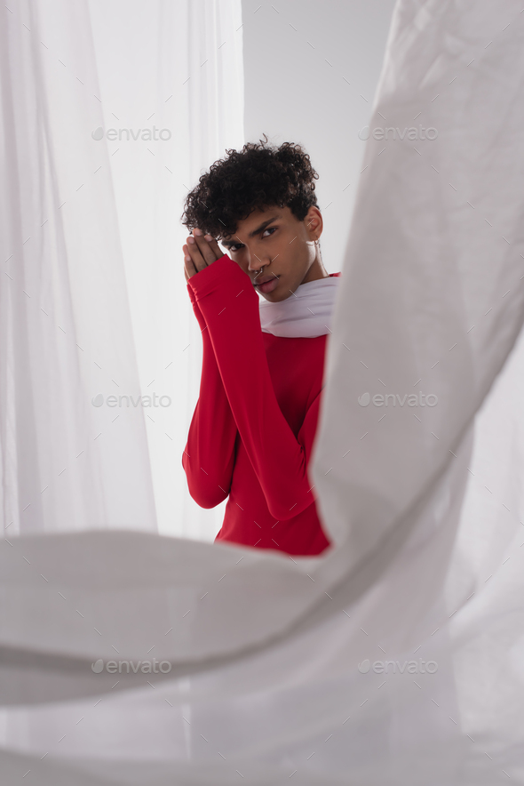 stylish african american man in red turtleneck posing with praying hands near white blurred drapery