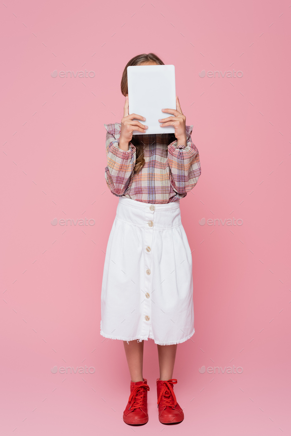 girl in white skirt, plaid blouse and red sneakers obscuring face with digital tablet on pink
