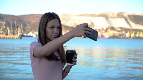 Attractive Young Woman Drinking Coffee and Using Mobile and Takes Selfies on Embankment with alt