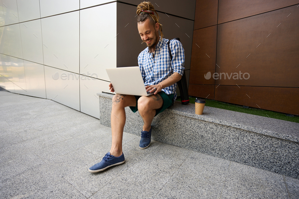 Happy freelancer chatting online on granite curb near modern building