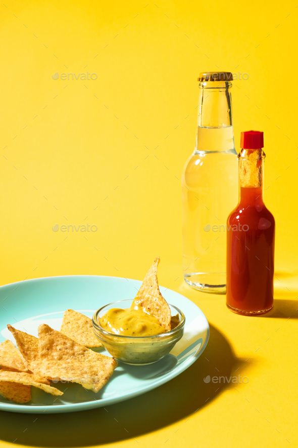 Nachos chips with cheese sauce on blue plate and soda bottle and hot sauce on yellow background.