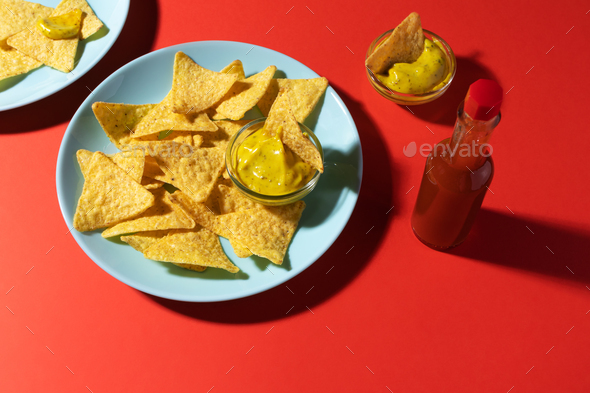 Nachos chips with cheese sauce on blue plate, soda bottle and hot sauce on red background