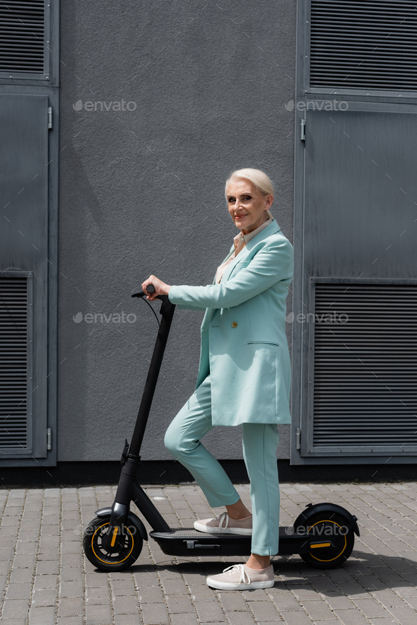 Smiling elderly businesswoman standing near electric kick scooter