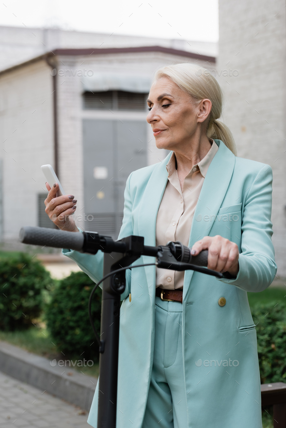 elderly businesswoman in blue blazer looking at mobile phone near electric kick scooter