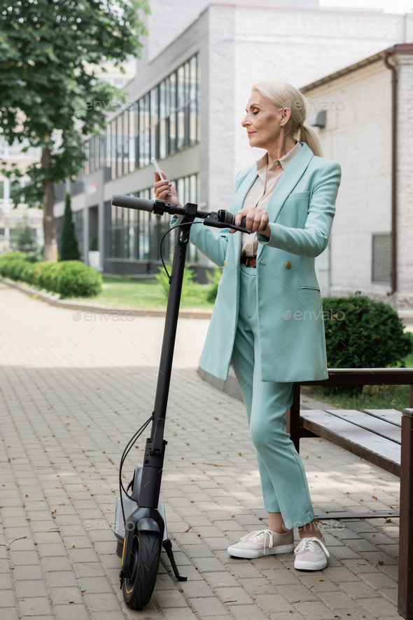 elderly businesswoman messaging on mobile phone near electric kick scooter