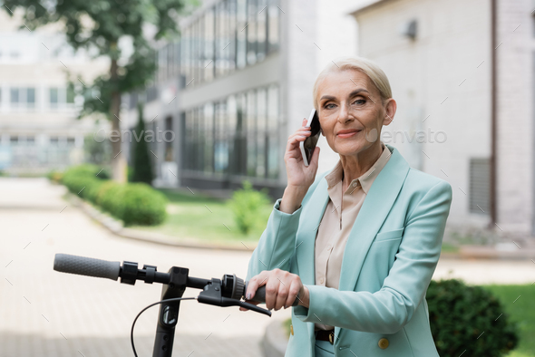 elderly businesswoman talking on mobile phone near electric kick scooter outdoors