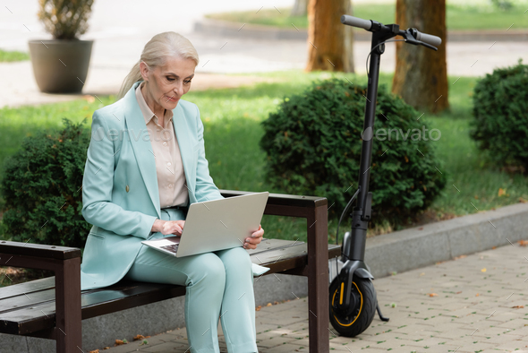 elderly woman using laptop on bench near electric kick scooter