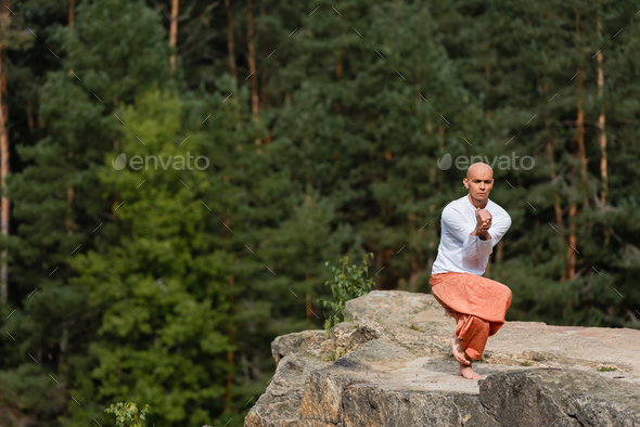 buddhist in white sweatshirt and harem pants meditating on rocky cliff in yoga pose