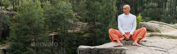 buddhist in white sweatshirt and harem pants meditating on rock in forest, banner