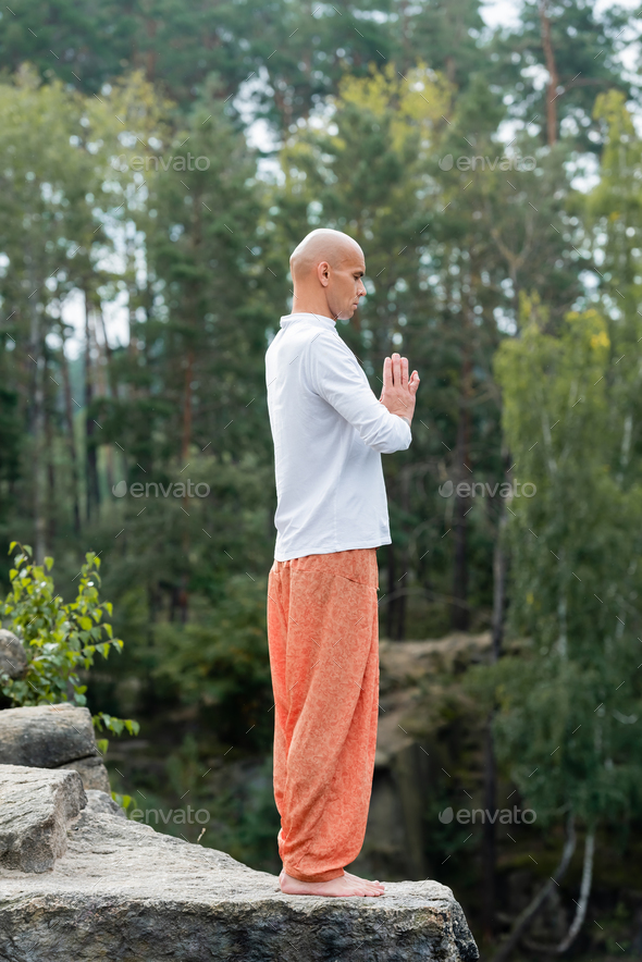 full length view of buddhist in sweatshirt and harem pants meditating with praying hands outdoors