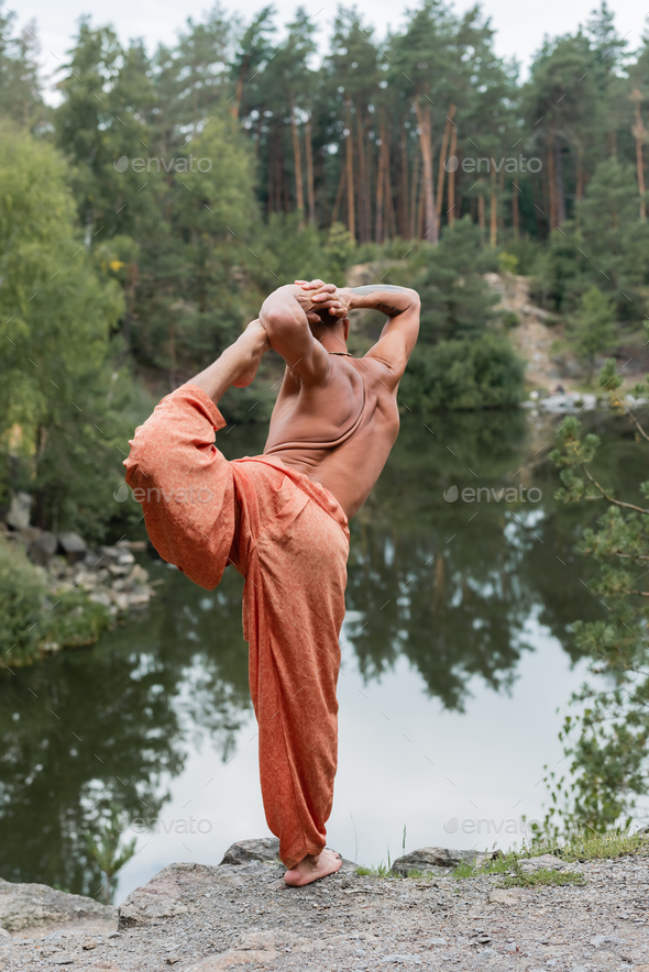 shirtless buddhist in harem pants practicing one legged bow pose on rocky cliff over forest lake