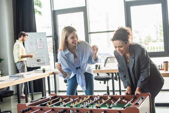 Excited businesswoman showing yes gesture near african american colleague playing table soccer in