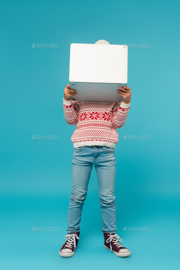 child in warm sweater and jeans obscuring face with laptop on blue