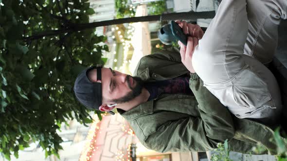 Young Man In Cap With Beard Sitting On The Street With Phone In His Hands alt