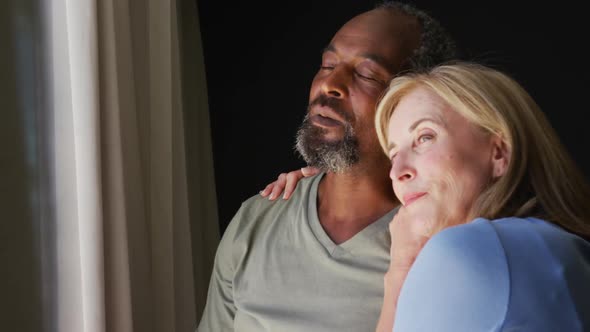Mixed race senior couple embracing each other while looking out of the window at home alt