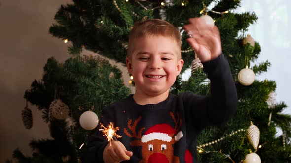 Little caucasian boy with Xmas sweater standing by Christmas tree at home and holding sparkler.